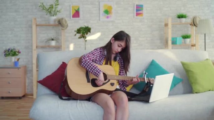 客厅中一个留着长发的美丽亚洲女孩的肖像，一个学习吉他的现代家庭在笔记本电脑上观看互联网上的教育视频。