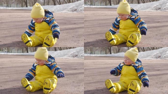 黄色防水雨裤围兜的孩子在寒冷的季节散步，一岁的男婴坐在地上玩耍。