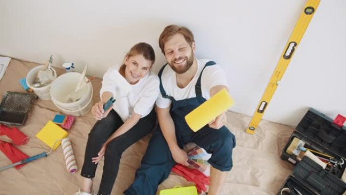 幸福的年轻夫妇在他们的新房子修理期间休息。与油漆滚筒一起坐在他们房子的地板上。年轻的夫妇坐在地板上选