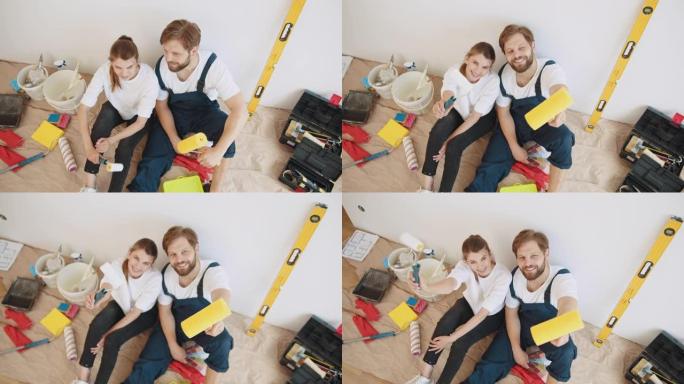 幸福的年轻夫妇在他们的新房子修理期间休息。与油漆滚筒一起坐在他们房子的地板上。年轻的夫妇坐在地板上选