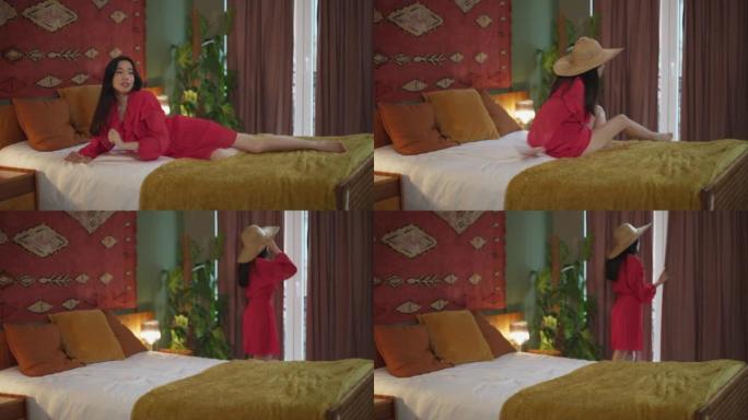 穿着红色夏装的美丽亚洲女人起床看窗户。女人不确定是否要在灰色的夏日出门-被困在酒店房间里