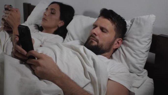 白人夫妇躺在床上睡觉前使用智能手机的特写镜头。男人和女人观看视频并浏览社交网络的提要