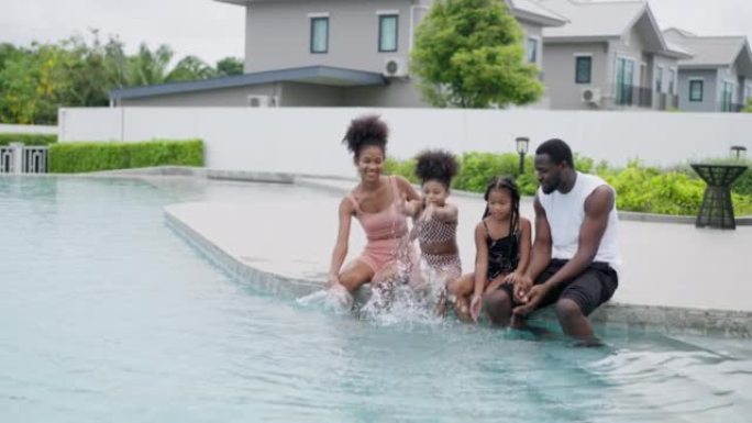 美国非裔家庭，肌肉发达的父亲，运动苗条的母亲和两个小孩正坐着，把腿放在水里。他们在游泳池边互相泼水，