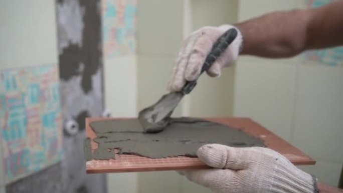 工作平铺机在涂胶之前，用带缺口的抹子将胶水涂在瓷砖上。男工师傅在浴室里墙上铺设瓷砖