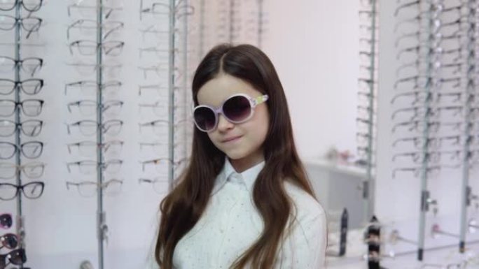 穿着可爱的白色开衫和衬衫的黑发女孩在眼镜店里戴着白边太阳镜，看着相机。眼科概念