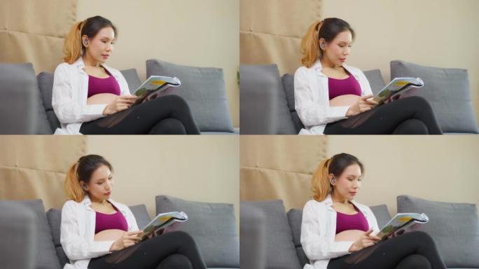 亚洲年轻女子孕妇坐在沙发上微笑看书。迷人的女性怀孕坐在沙发上寻找指南，为子宫中的婴儿做准备，在客厅里