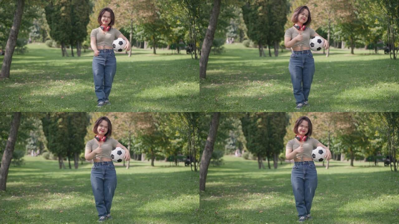 宽镜头适合侏儒症的年轻女子，指着拇指微笑着看着夏天公园里拿着足球站着的相机。正面小人物慢动作摆姿势。