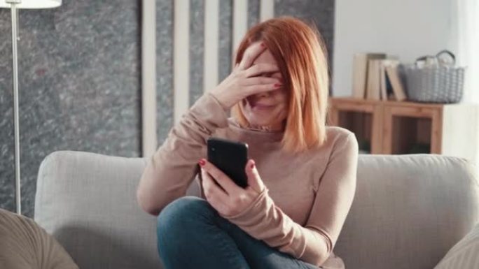 成年妇女坐在沙发上拿着手机阅读不愉快的消息坏短信感到沮丧开始哭泣。