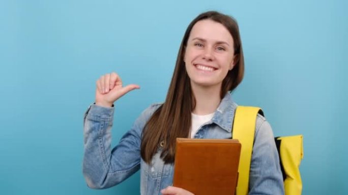 开朗可爱的女孩青少年学生穿着牛仔夹克和黄色背包，用手指指着自己的书，在蓝色工作室背景下孤立地摆姿势。