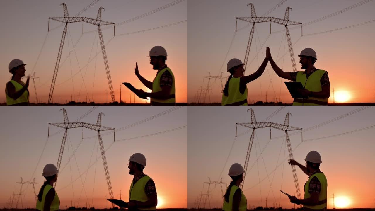两名电气工程师在日落时高压电塔的背景下击掌。电网附近工人会议。团队合作，签订合同，两个商人