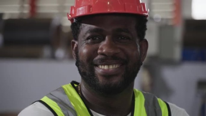 在工厂工业中，快乐的职业非裔美国男子工人的肖像戴着安全帽站着微笑，看着相机。