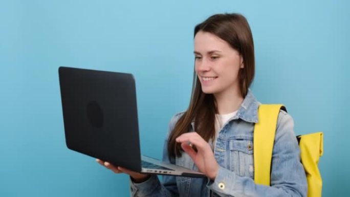肖像漂亮女孩青少年学生穿牛仔夹克和黄色背包在笔记本电脑上保持使用类型的作品，显示拇指向上，孤立的蓝色