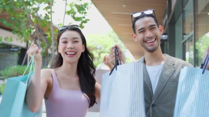 在百货商店户外购物的亚洲男女肖像。迷人的年轻夫妇拿着购物袋，然后幸福地散步，享受在购物中心购物和看相