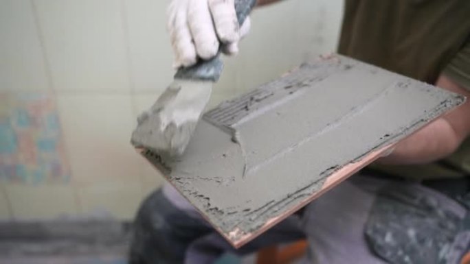 工作平铺机在涂胶之前，用带缺口的抹子将胶水涂在瓷砖上。男工师傅在浴室里墙上铺设瓷砖