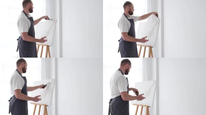 一位年轻的欧洲大胡子艺术家在干净的白色画布上涂上白色油漆