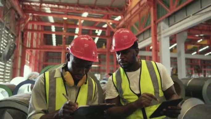 两名身穿制服的非洲男子工人戴着安全帽，拿着平板电脑和剪贴板检查工厂库存。