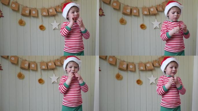 穿着睡衣的男孩，圣诞老人帽子在带有礼物背景的纸质降临日历上吃甜糖果。墙上有手工圣诞日历礼物的小快乐孩
