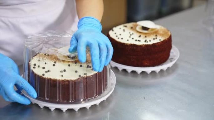桌上摆着两个装饰精美的蛋糕。工人戴着手套的手用塑料盖覆盖甜点。特写。
