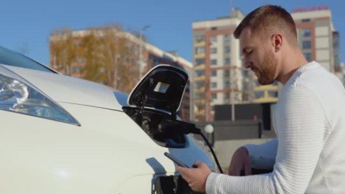 皮肤白皙的男司机将电动汽车连接到电源系统，为汽车电池充电，并通过智能手机控制充电过程