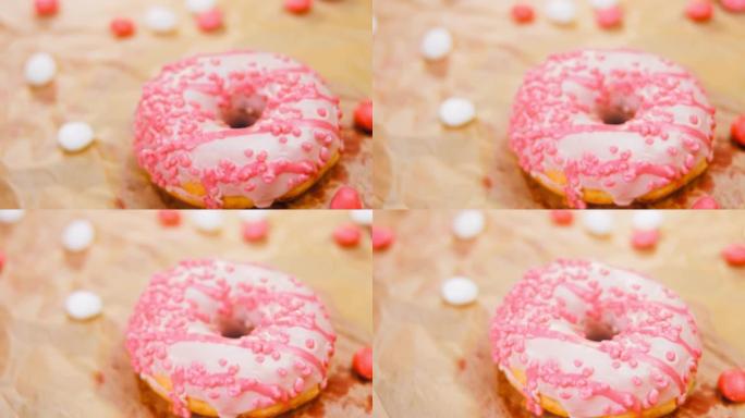粉色甜甜圈装饰有粉色糖衣。甜甜圈在一张用糖果装饰的纸上。微距和滑块拍摄。糖果以慢动作移动。面包店和食