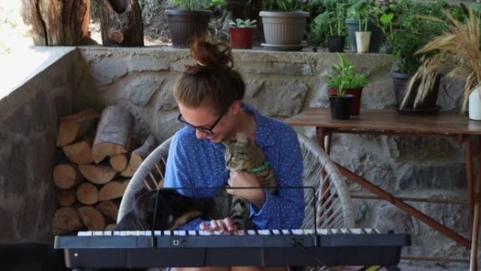 戴着眼镜的年轻女子在乡下的露台上玩合成器时，和她的猫和狗玩得很开心。
