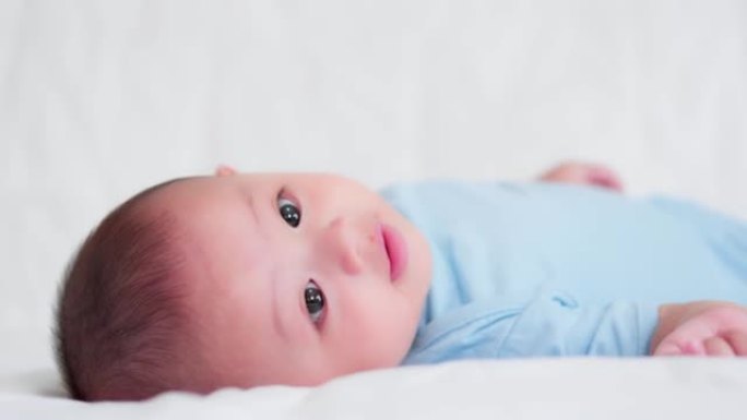 幸福的家庭，可爱的亚洲新生婴儿穿着蓝色衬衫躺在白色床上玩耍，看着相机笑着开心的脸。天真的小新婴儿可爱