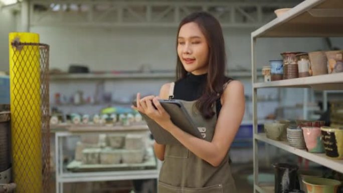 亚洲女陶艺家用围裙手将她的粘土雕刻产品放在架子上等待烤箱烘烤，亚洲女小企业陶瓷车间老板微笑着自豪地展