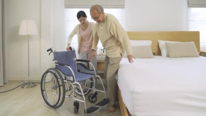 亚洲女护士，女儿帮助父亲从床上爬到轮椅上，在家里的卧室或医疗保健的房子里为老年老年患者提供支持。人们