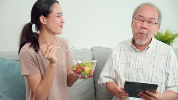 照顾者年轻的亚洲妇女喂养蔬菜老年亚洲男子坐在家里的客厅沙发上。女治疗师正在监测老年男性患者的症状。医