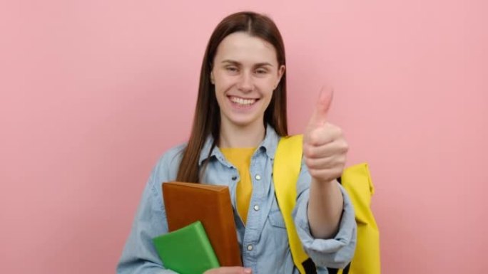 微笑的漂亮女孩青少年学生拿着书指指相机你露出拇指，穿着衬衫和黄色背包，在粉色背景工作室中孤立地摆姿势