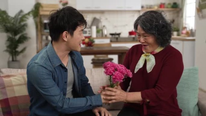 韩国成年儿子的慢动作在家里的客厅沙发上给奶奶惊喜的康乃馨花。微笑的退休女性说哇，并为母亲节概念幸福地