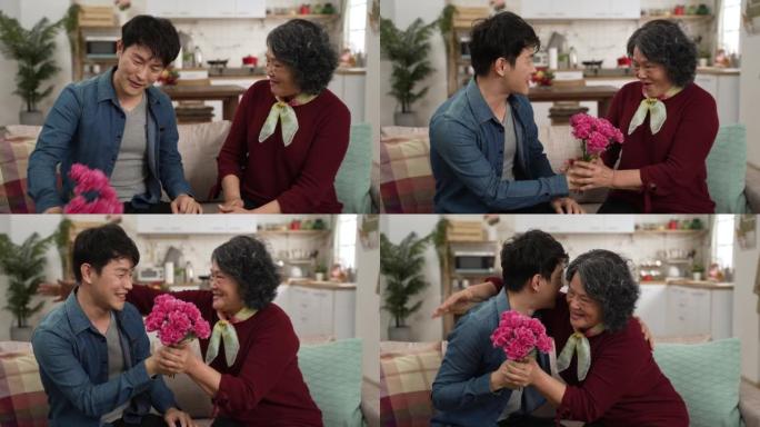 韩国成年儿子的慢动作在家里的客厅沙发上给奶奶惊喜的康乃馨花。微笑的退休女性说哇，并为母亲节概念幸福地