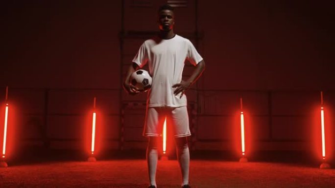 运动黑人男子手持足球，晚上独自站在足球场上，艺术红灯