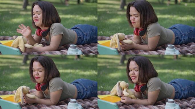 放松积极的小女人在春夏公园里躺在毯子上玩玩具。快乐快乐的高加索人的肖像，侏儒症，享受户外休闲。慢动作