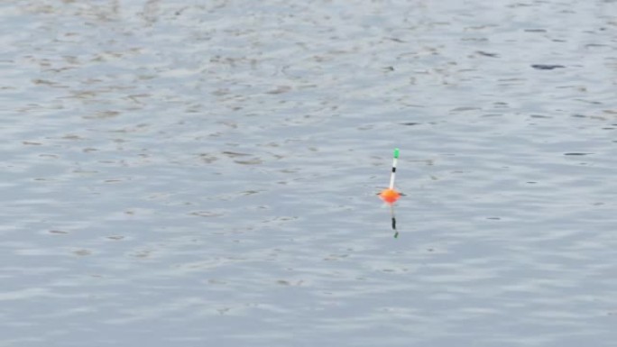 一个红色和绿色的钓鱼浮标在海浪上摆动，同时在河上用钓鱼竿钓鱼的特写镜头。