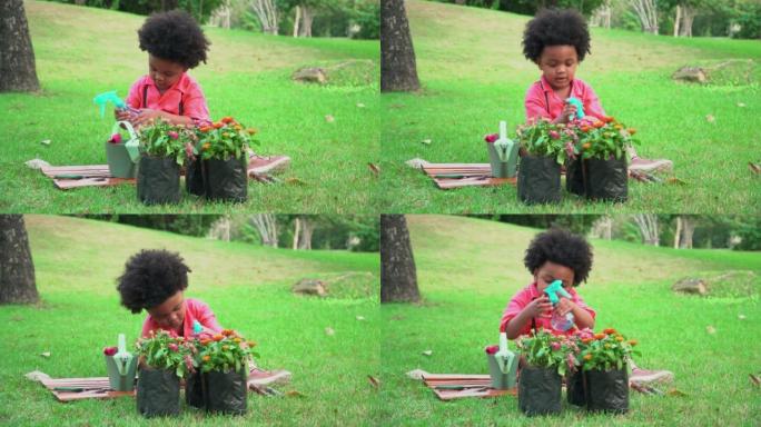 可爱的美国非洲裔小男孩坐在绿草环绕的垫子上，正在用喷壶浇花树，并改用喷雾瓶向公园的小花喷洒水滴。