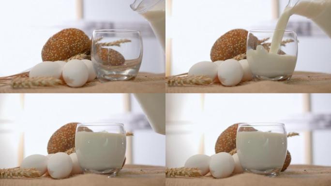 将新鲜的牛奶倒入质朴的稻草桌上的玻璃杯中，将白色鸡蛋与小麦豆荚和一杯牛奶的特写镜头。食物静货