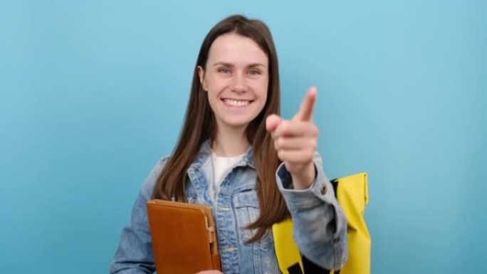 肖像快乐女孩青少年学生拿着书眨眼眨眼，穿着牛仔夹克和黄色背包，孤立在工作室的蓝色背景上。高中教育大学
