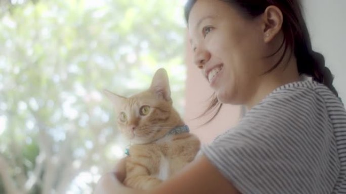 亚洲女人抱着虎斑猫看着新家玻璃窗边的景色。收养流浪猫的概念要有一个爱并能照顾他们的新主人。