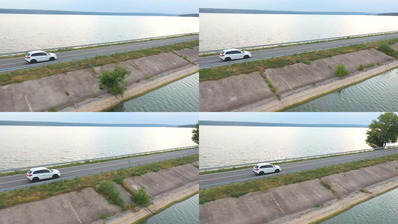 夏日白色汽车穿越大坝路线的空中拍摄。SUV在靠近湖泊或池塘的道路上行驶。飞越汽车穿过河桥。周末旅行或