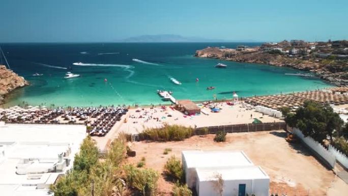 在晴朗的晴天，希腊米科诺斯岛的超级天堂海滩拍摄了空中无人机轨道，山上有游客和水上运动居民。