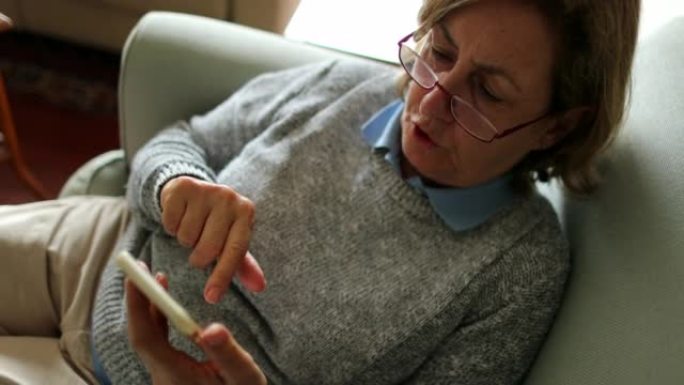 老年妇女坐在家里的沙发上使用手机，手持智能手机的老年人
