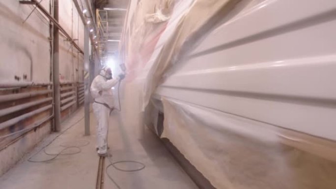 一名穿着防护服和呼吸器的男子使用粉碎机用清漆保护层覆盖了汽车。在油漆车间的马车厂工作