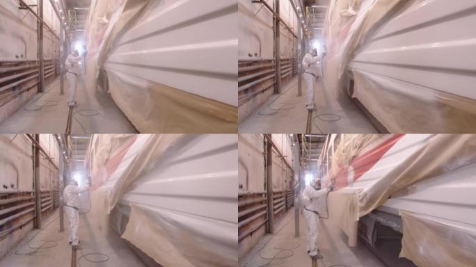 一名穿着防护服和呼吸器的男子使用粉碎机用清漆保护层覆盖了汽车。在油漆车间的马车厂工作