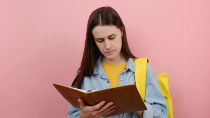 20多岁的少女学生肖像穿着衬衫和黄色背包阅读书籍《思考梦想》，在工作室的粉色背景墙上孤立地摆姿势。高
