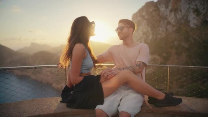 浪漫的夏天。夫妇在度假时戴着墨镜坐在长凳上，背景是金色的日落。女人在温暖的怀抱中躺在男人的腿上