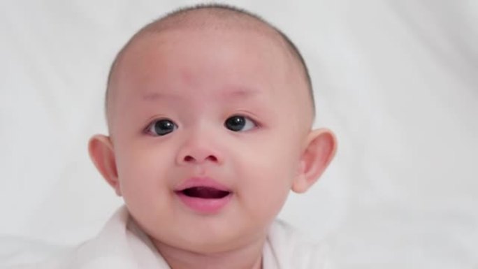 脸部特写可爱的亚洲新生婴儿躺在白色床上玩耍看着相机笑着开心的脸。天真的小新婴儿可爱。