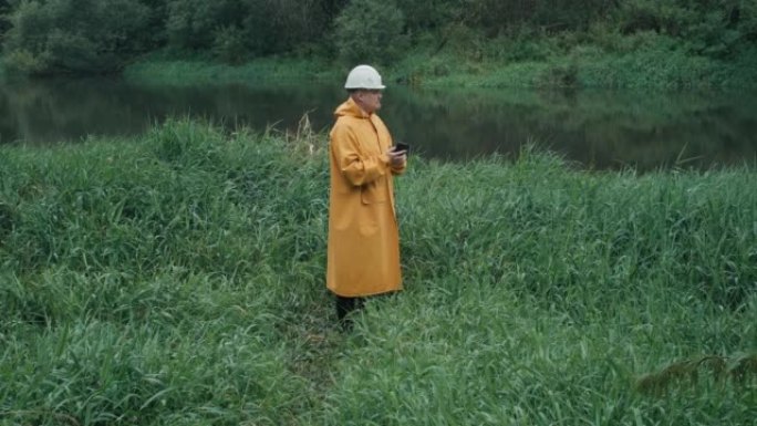 戴着头盔和雨衣的工人走到草地上的河边，男人手里拿着电话