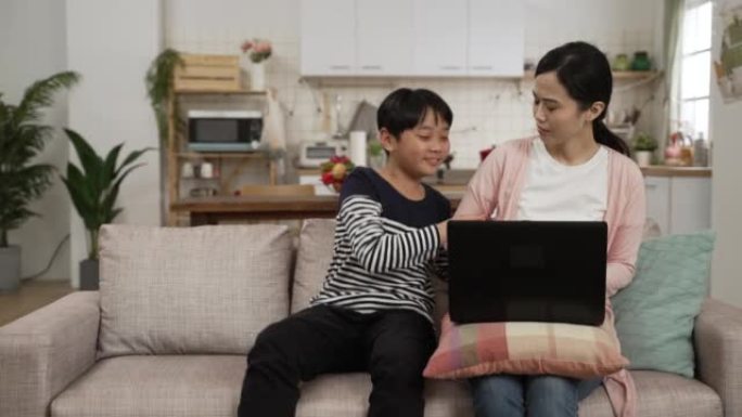 忙碌的亚洲日本妈妈正在用笔记本电脑工作。淘气的儿子在家里的客厅里跑来跑去和他唠叨