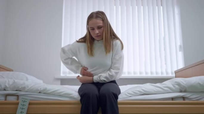 悲伤疲惫的年轻女子，严重的胃痛坐在医院病房的床上。在室内诊所等待医生的高加索患者的正面肖像。阑尾炎炎